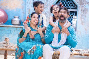 Cheerful rural indian family sitting on traditional bed at village home, excited little kids having fun with parents. daughter and son hug father and mother. happy life, playful children.