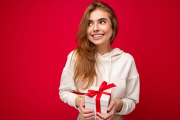 Shot of attractive positive smiling young blonde woman isolated over colourful background wall wearing everyday trendy outfit holding gift box and looking to the side