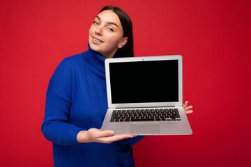 Charming pretty happy beautiful young brunet female student lady holding computer laptop looking at camera in blue sweater isolated over red wall background