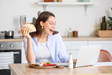 Beautiful young woman with laptop eating tasty quesadilla at home