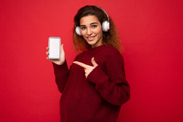 Shot of attractive positive young brunet curly woman wearing dark red sweater isolated on red background wall wearing white wireless headphones and listening to music and showing mobile phone with