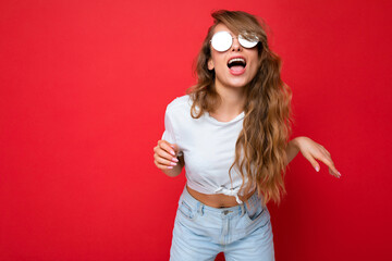 Shot of beautiful amusing joyful young dark blonde curly woman isolated over red background wall wearing casual white t-shirt and stylish sunglasses looking at camera