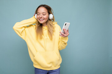 Naklejka premium Photo shot of beautiful joyful smiling young female person wearing stylish casual outfit isolated over colorful background wall wearing white bluetooth wireless earphones and listening to music and