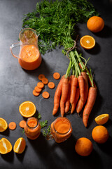 bunch of organic carrots, with their green stems, loose carrot slices, oranges and a jug with natural juice and two glasses with carrot juice on a gray background,