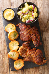 Authentic Schwenkbraten Marinated BBQ Pork neck steak with potatoes salad, mustard and toast close up in the slate board on the table. Vertical top view from above