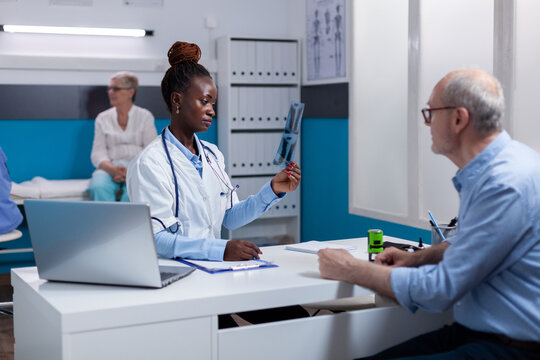 Medic Of African American Ethnicity Holding X Ray For Old Patient Checkup In Medical Cabinet. Black Doctor With Radiography Explaining Diagnosis To Elderly Sick Person Sitting At Desk