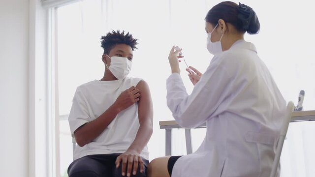African American teenager wearing face mask sit in hospital waiting for COVID-19 vaccination. Epidemic Control with Inject at Clinic. COVID-19 Vaccination station. Free quality vaccines for masses.