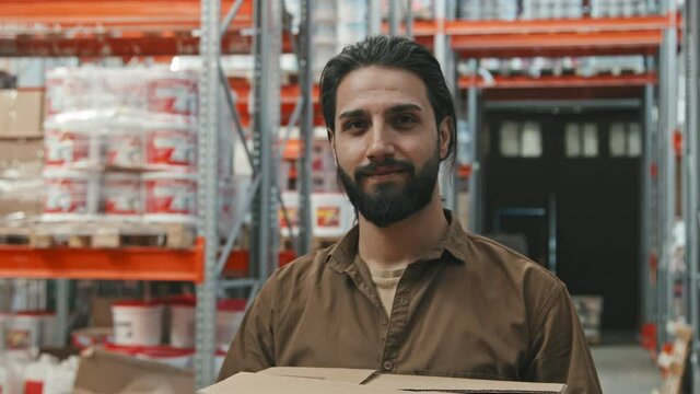 Slowmo tracking portrait shot of young man with beard holding boxes and smiling for camera at hardware store warehouse
