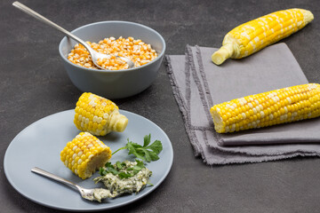 Two boiled ears of corn on gray napkin. Pieces of boiled corn, sauce and spoon on gray plate.