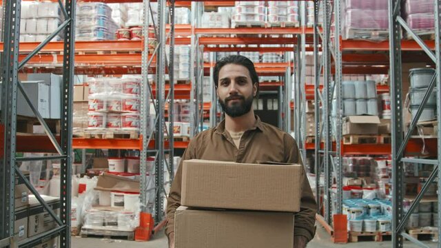 Slowmo tracking portrait of bearded young male worker holding cardboard boxes and posing for camera in hardware store warehouse