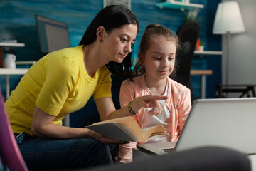 Caucasian mother helping girl with homework project at home. Parent assisting child while reading book for education exam and online class lesson lecture with modern laptop at desk