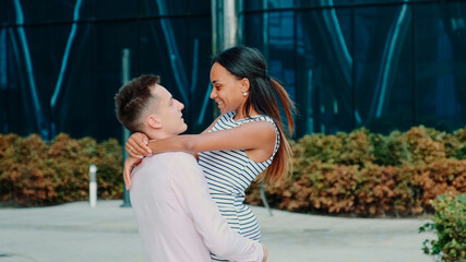 Smiling multiracial couple having fun spinning around on the street. A big office building is in the background.