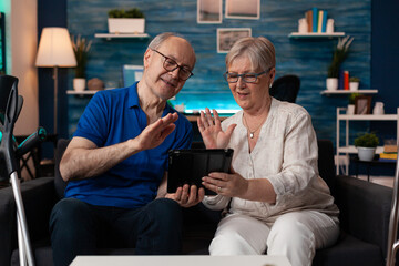 Old husband and wife waving on video call using tablet with online internet technology gadget sitting at home. Caucasian couple having virtual conference connection for communication