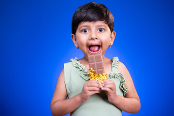 Excited Girl kid with large eyes open, enjoying eating chocolate on blue studio background -...
