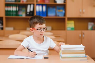 Concentrated schoolboy sitting at desk and writing in exercise book with classmate sitting behind