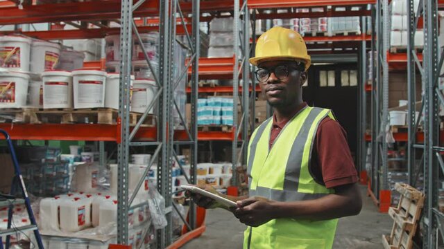 Slowmo tracking portrait of African-American male worker in hard hat and safety vest using tablet in warehouse of hardware store, then looking at camera