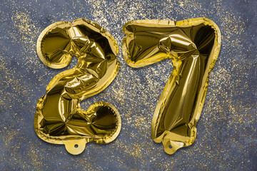 The number of the balloon made of golden foil, the number twenty-seven on a gray background with sequins. Birthday greeting card with inscription 27. Numerical digit, Celebration event, template.