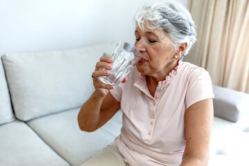 Beautiful old woman drinking water. Happy senior woman with glass of water at home. Thirsty woman drinking a refreshing healthy glass of cold fresh water. Age, health care and people concept.