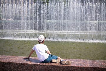 Boy sitting on fence of fountain and looking at the water, wearing white T-shirt and cap, view from the back