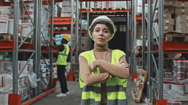 Handheld slowmo tracking of female warehouse worker in hard hat and reflective vest looking away from camera, then turning and posing with her arms crossed