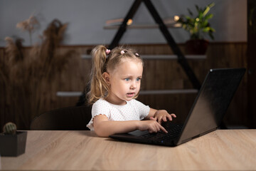 One little preschool girl in the evening sits at home at a table with a laptop.