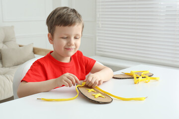 Little boy tying shoe lace using training cardboard template at white table indoors - obrazy, fototapety, plakaty