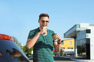 Young man with coffee eating hot dog near car at gas station