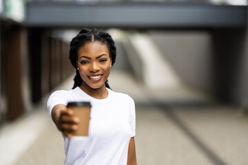 Cheerful young african woman walking outdoors, holding takeaway coffee cup and pointed on you