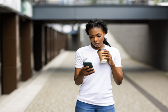 Cheerful young african woman holding takeaway coffee cup and using mobile phone while walking outdoors. 