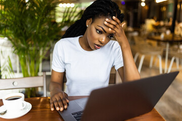 Pretty African-American woman sitting at table near laptop and looking at camera with tired face expression.