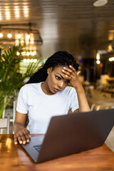 Pretty African-American woman sitting at table near laptop and looking at camera with tired face expression.