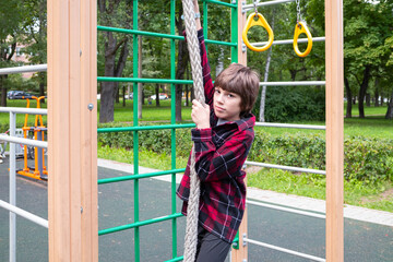 Boy in red plaid shirt playing on crossbars in playground in summer on background of green trees