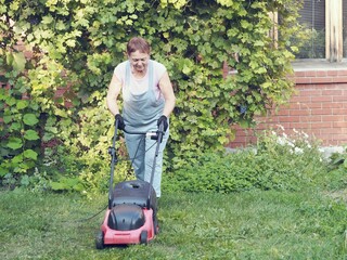 Senior woman mows a green lawn with a pushing electric mower.