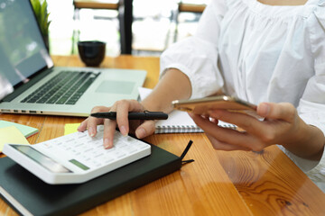 Businesswoman calculating with calculator, holding smartphone, laptop on desk