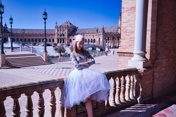 Adult female Hispanic classical ballet dancer in white tutu and pink cancer scarf with her arms crossed and looking serious leaning on a stone railing.