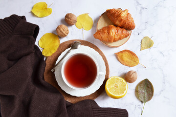 Cup of tea with falling yellow leaves, croissants, lemon and walnuts. Hello autumn. Tea break at home. Morning tea routine. English morning concept.  Thanksgiving holiday composition. Warm cozy home.