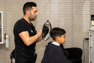 Fototapeta premium Barber shows the result of a haircut to a child with a mirror. Boy checks through the mirror how his haircut has turned out.