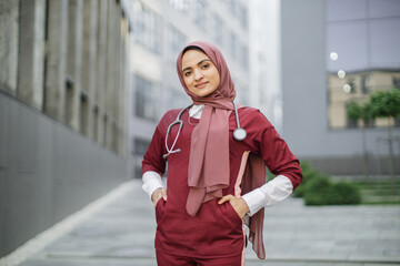 Beautiful Malay lady doctor wearing hijab, scrubs and stethoscope outside modern clinic. Closeup...