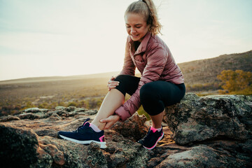 Caucasian female hiking on mountain holding sprained ankle in pain waiting for help sitting on rock - obrazy, fototapety, plakaty