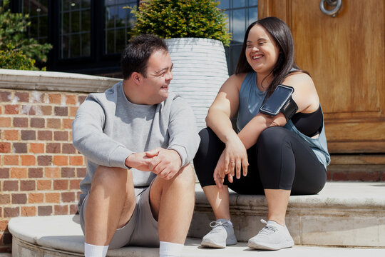 Young biracial couple with Down Syndrome in active wear and wearable tech sitting on steps and laughing