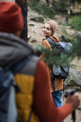 happy woman with backpack looking at man trekking in forest on blurred foreground