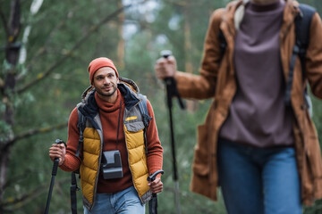 man with vintage camera looking at woman holding hiking sticks while trekking in forest on blurred foreground
