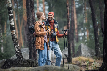 cheerful couple in hats holding hiking sticks while hugging in forest