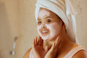 Young biracial woman with Down Syndrome applying face mask in the bathroom and smiling