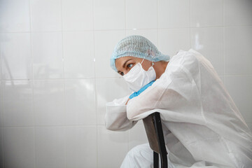 A young female doctor in a protective suit, cap, mask and gloves is sitting tired on a chair in the medical office