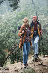 full length of couple in hats holding hiking sticks while trekking in forest