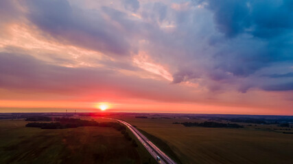 Aerial view of highway on red sunset. Landscape with road near countryside fields. Beautiful winding road leading through rural countryside with evening sunlight. Dramatic sky background.