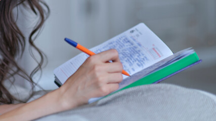 lifestyle soft image of unrecognizable young woman sitting on her cozy bed and making notes to her diary. Fall season mood.