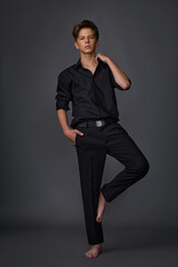 guy teenager in a black shirt and black trousers posing barefoot in the studio on a gray background