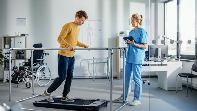 Hospital Physical Therapy Room: Patient with Injury Walking on Treadmill Holding for Parallel Bars, Professional Physiotherapist Assists, Helps, Trains Disabled Person Do Rehabilitative Physiotherapy 
